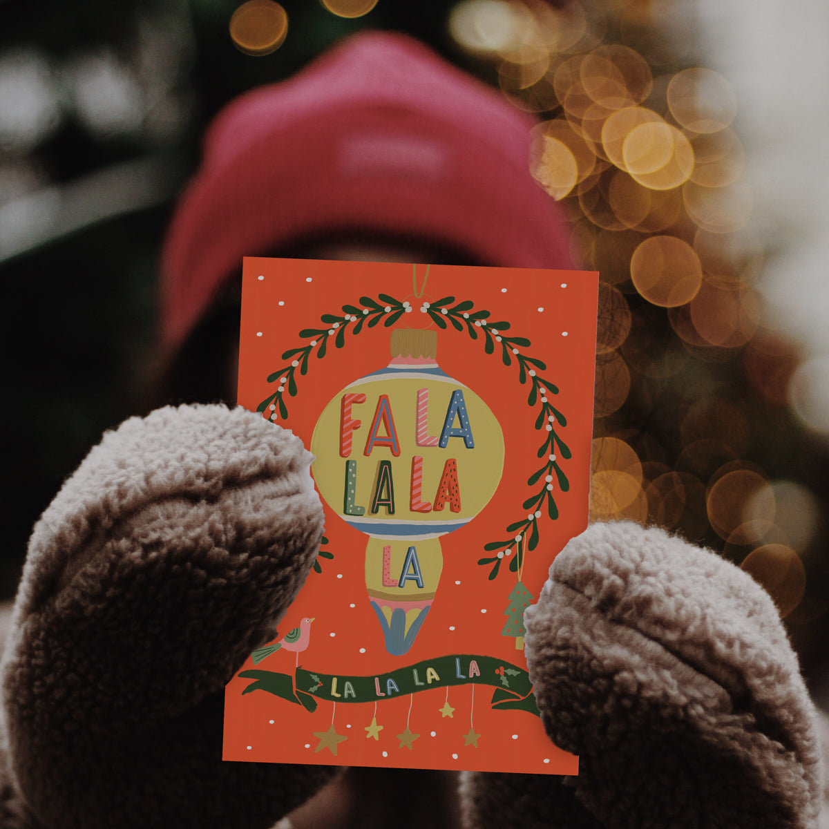 Christmas Card Fa la la with mistletoe, dove Christmas tree and bauble. Illustrated by Studio Peers. Mock details girl holding a Festive Card red and gold.